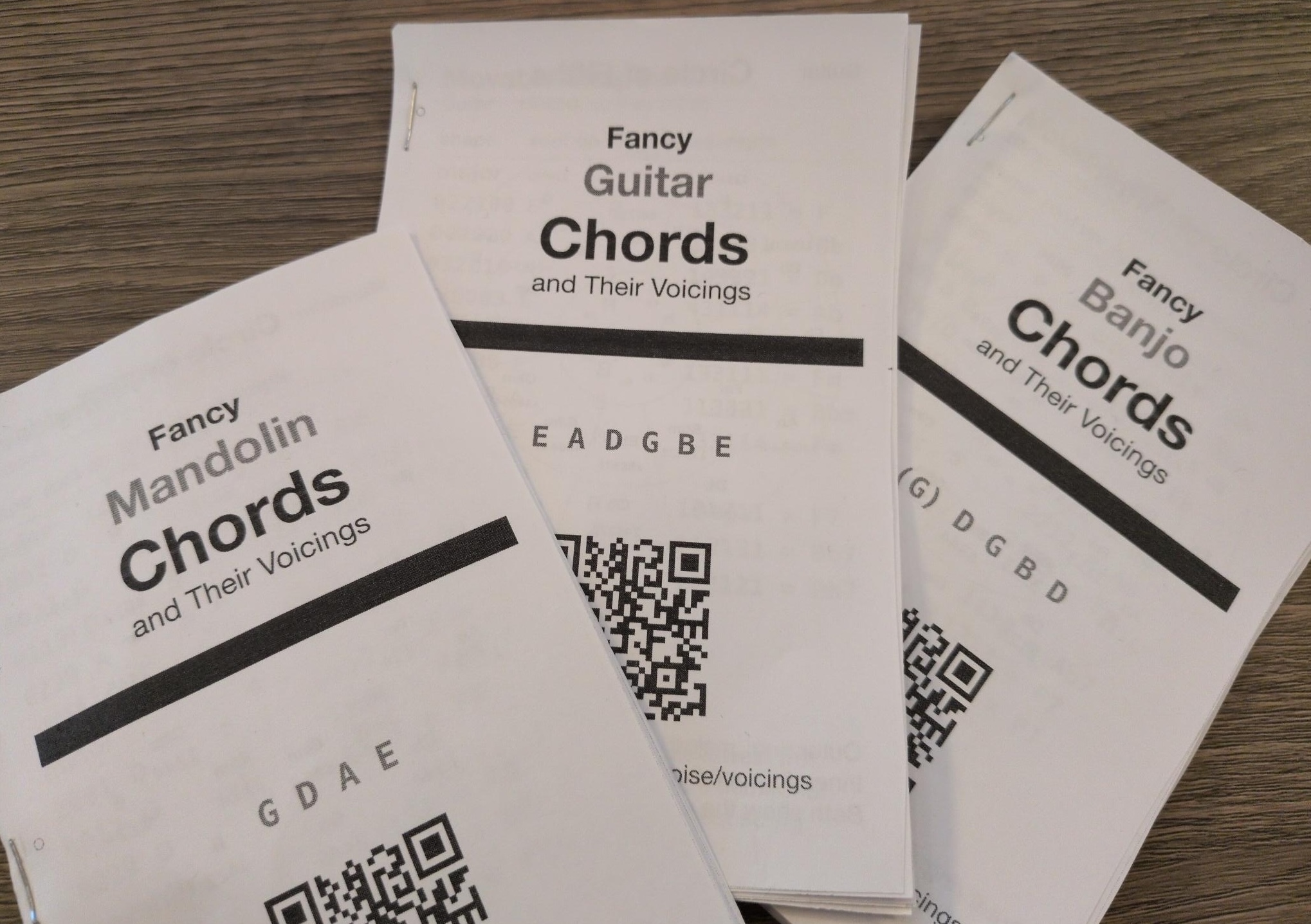Three stapled
booklets on a desk: Fancy Mandolin Chords, Fancy Guitar Chords and Fancy
Banjo Chords, each with its tuning and a QR code on the cover